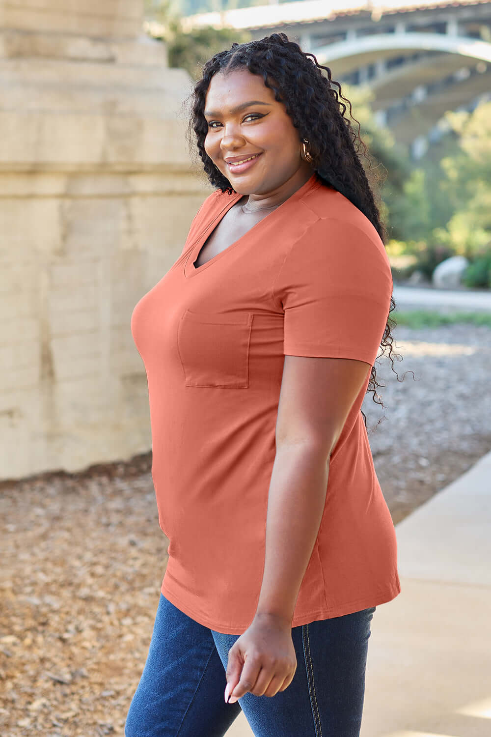 Woman wearing Basic Bae Bamboo V-Neck Short Sleeve T-Shirt in orange, smiling outdoors while showcasing the comfy and stylish tee.