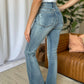 Woman wearing Judy Blue medium rise tummy control flare jeans with distressed detail, standing in stylish living room setting.