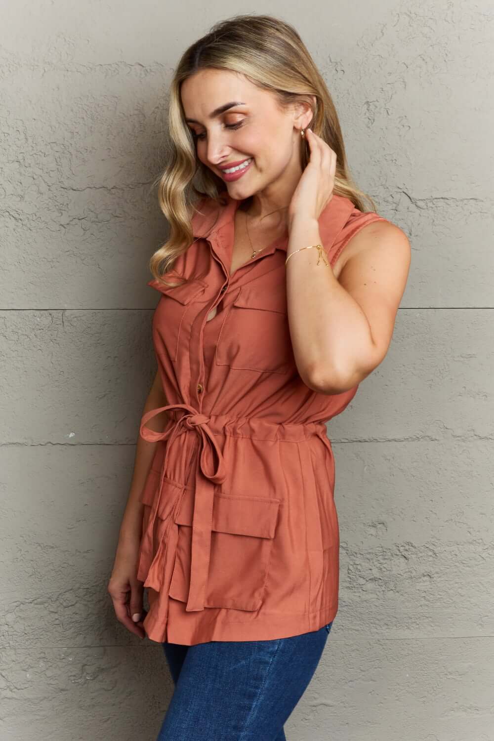 Woman wearing Follow The Light Sleeveless Collared Button Down Top in rust color with button-down front and tied waist, paired with blue jeans.