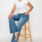 Model showcasing bytos Cat's Whiskers Mid Rise Bootcut Jeans with a stylish white top and heels, sitting on a stool.