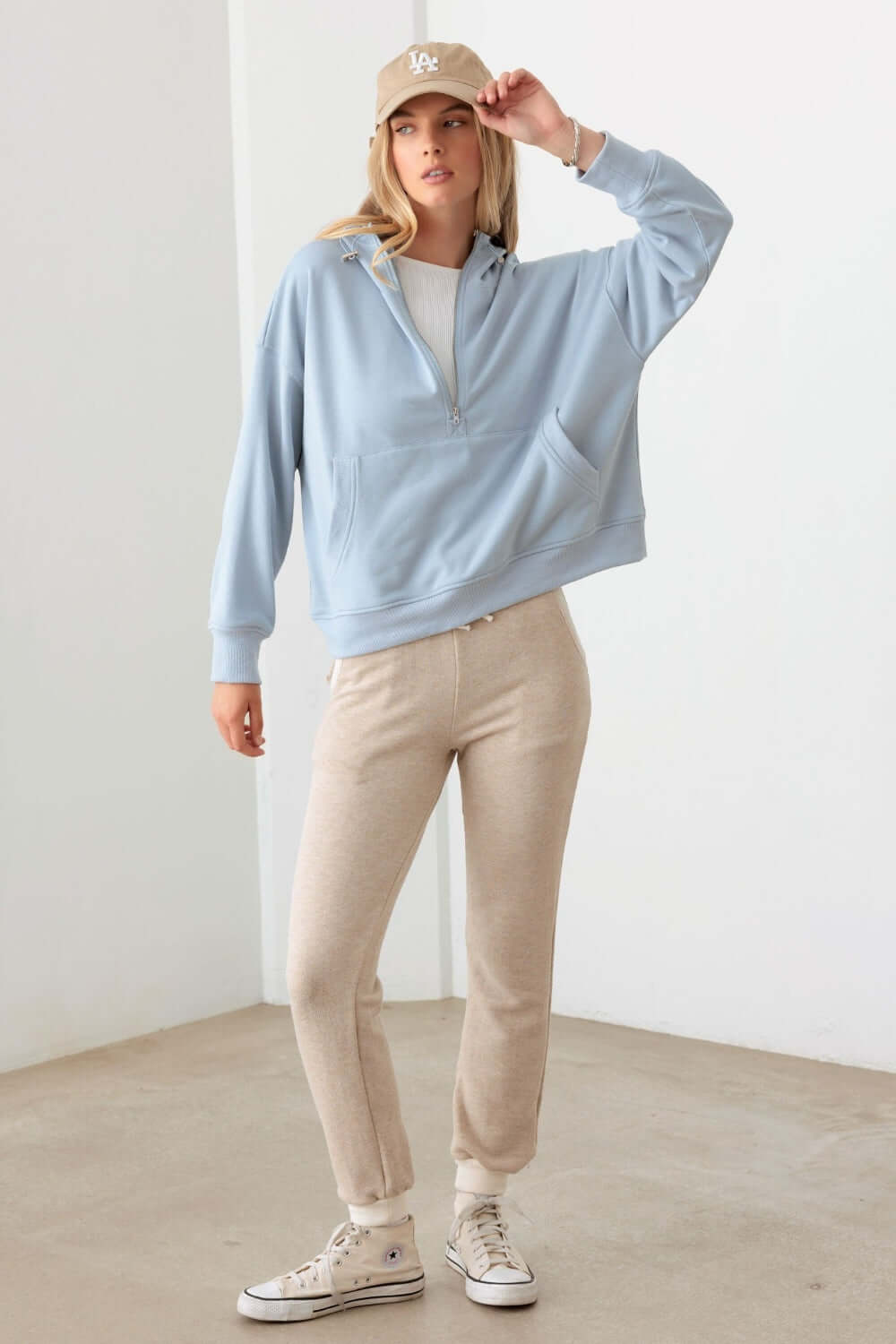 Woman wearing a Tasha Apparel Half Zip Drawstring Mock Neck Hoodie in blue with beige sweatpants and sneakers, posing against a white background.