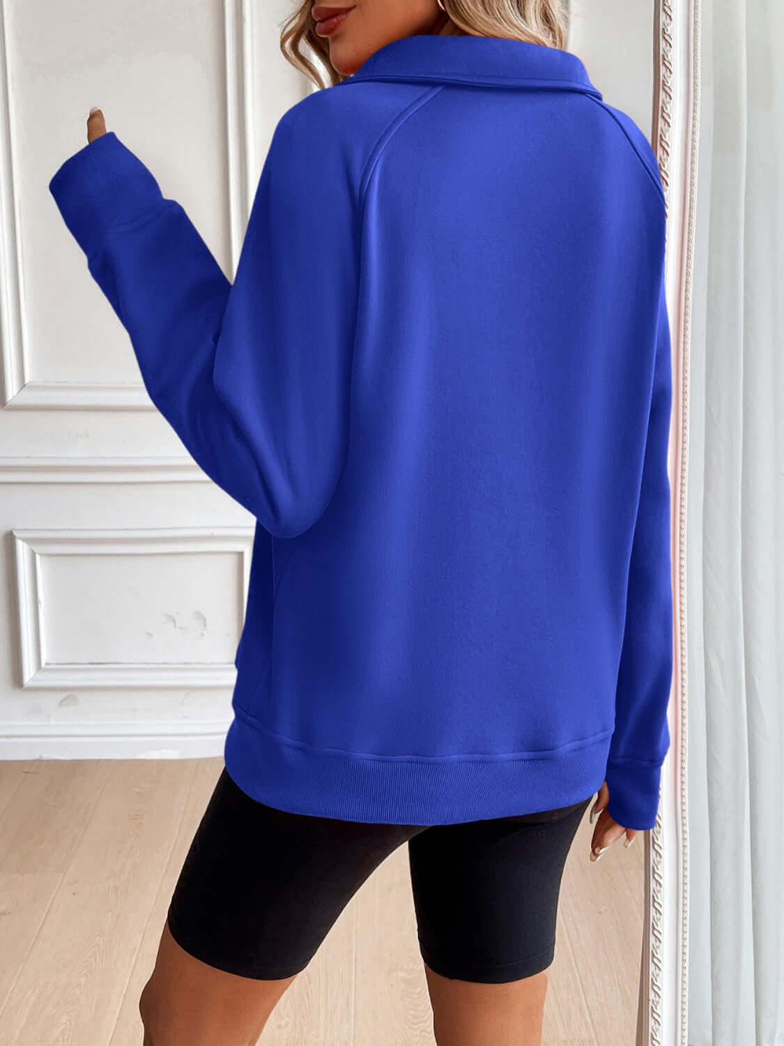 Woman wearing Ivy Lane Half Zip Raglan Sleeve Sweatshirt in blue, viewed from the back with her hand touching the collar in a stylish setting.