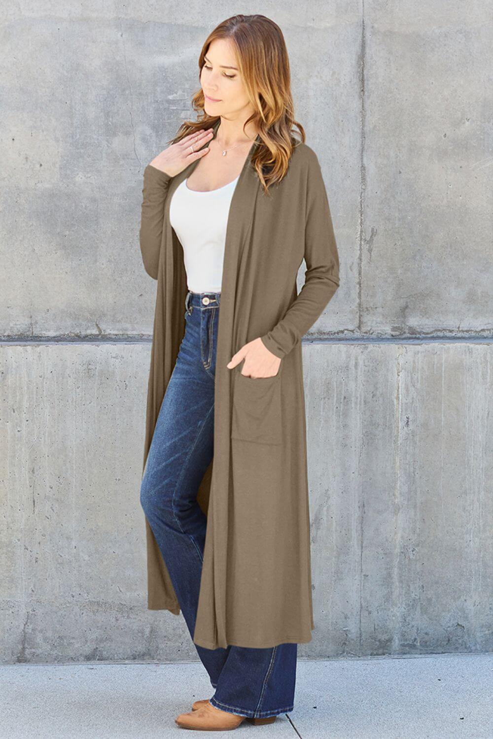 Woman wearing open front long sleeve cover up with pockets, styled with white top and blue jeans, standing against concrete wall