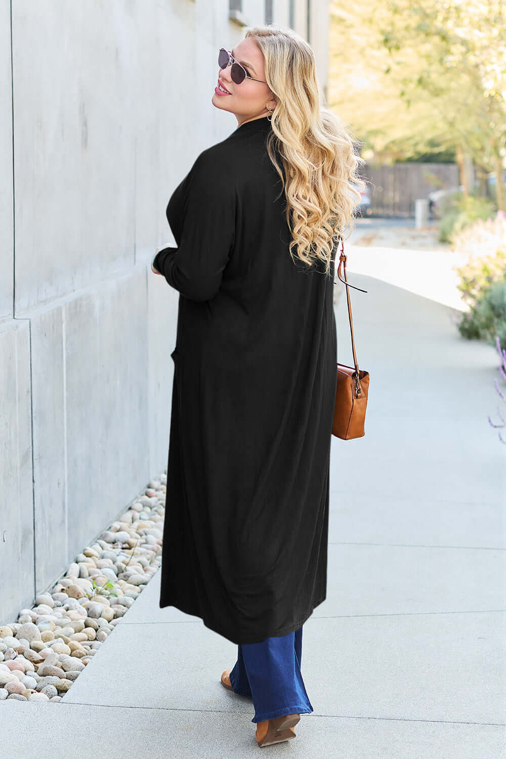 Woman modeling black open front long sleeve cover up with pockets, outdoors, wearing sunglasses and holding a brown bag.