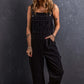 Woman wearing black pocketed straight denim overalls standing against a dark textured background.