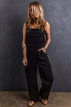 Woman wearing black pocketed straight denim overalls standing against a dark textured background.