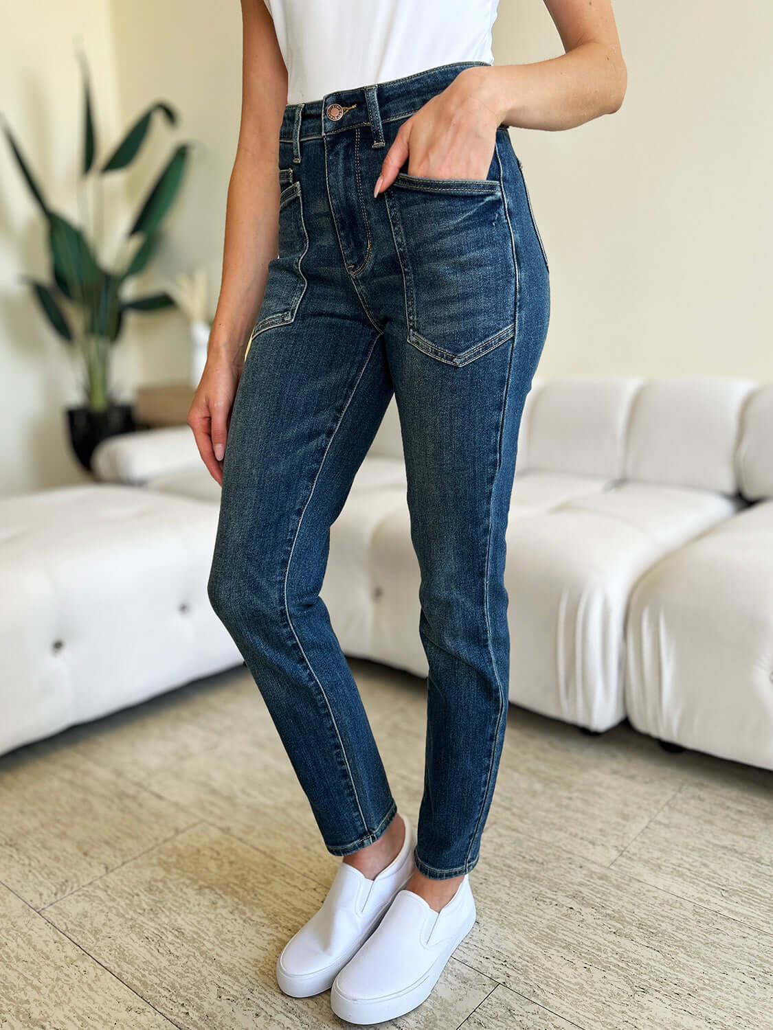 Woman in High Waist Skinny Judy Blue Jeans with white sneakers standing in a modern living room.