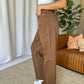 Woman wearing RFM Full Size High Rise Garment Dye Wide Leg Jeans in brown, standing in a stylish living room with a sofa in the background.