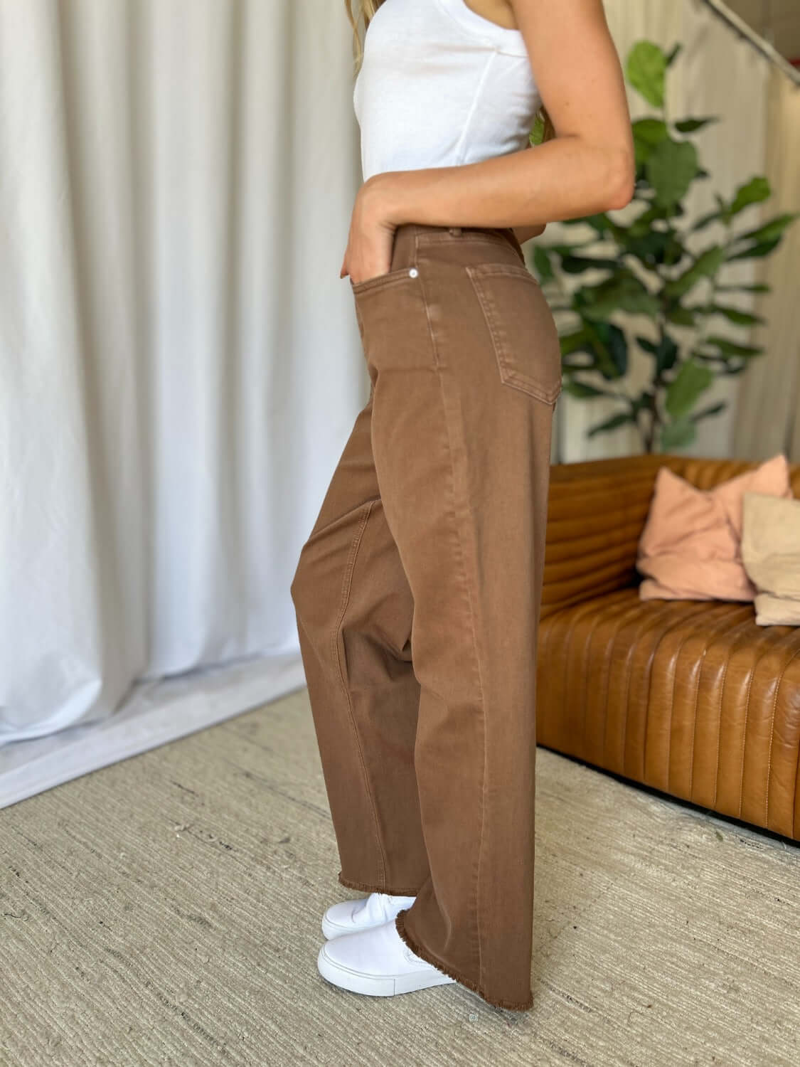 Woman wearing RFM Full Size High Rise Garment Dye Wide Leg Jeans in brown, standing in a stylish living room with a sofa in the background.