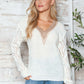 Woman wearing Bella Road Openwork V-Neck Long Sleeve Sweater in white with stylish design, standing against rustic wooden wall.