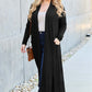 Woman wearing black open front long sleeve cover up over white top and blue jeans, accessorized with sunglasses and crossbody bag, walking