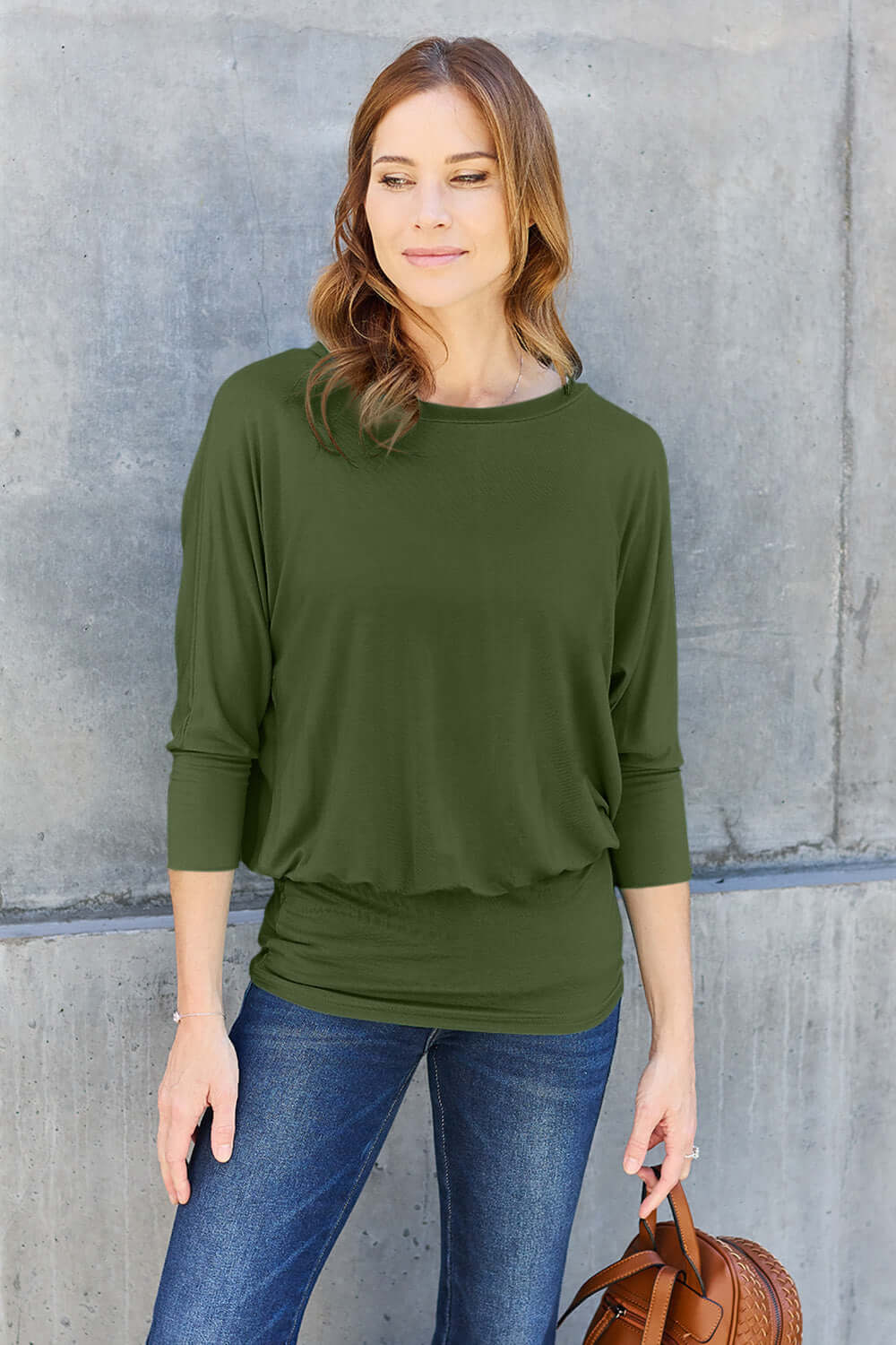 Woman wearing green round neck batwing sleeve top with jeans against concrete wall.