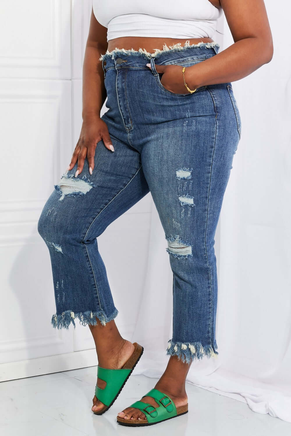 Woman wearing Undone Chic Straight Leg Risen Jeans with distressing, raw hems, and waist details paired with a white crop top and green sandals.