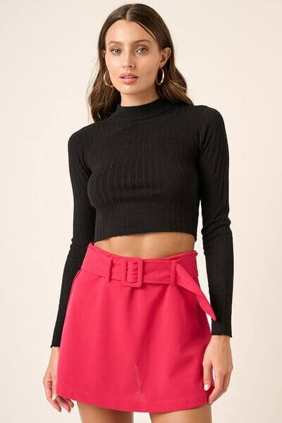 Model wearing a black mock neck long sleeve crop knit top paired with a pink belted mini skirt, showcasing a chic outfit.