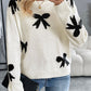 Woman wearing a Perfee Bow Graphic Round Neck Long Sleeve Sweater in white with black bow designs, paired with blue jeans and a black clutch.