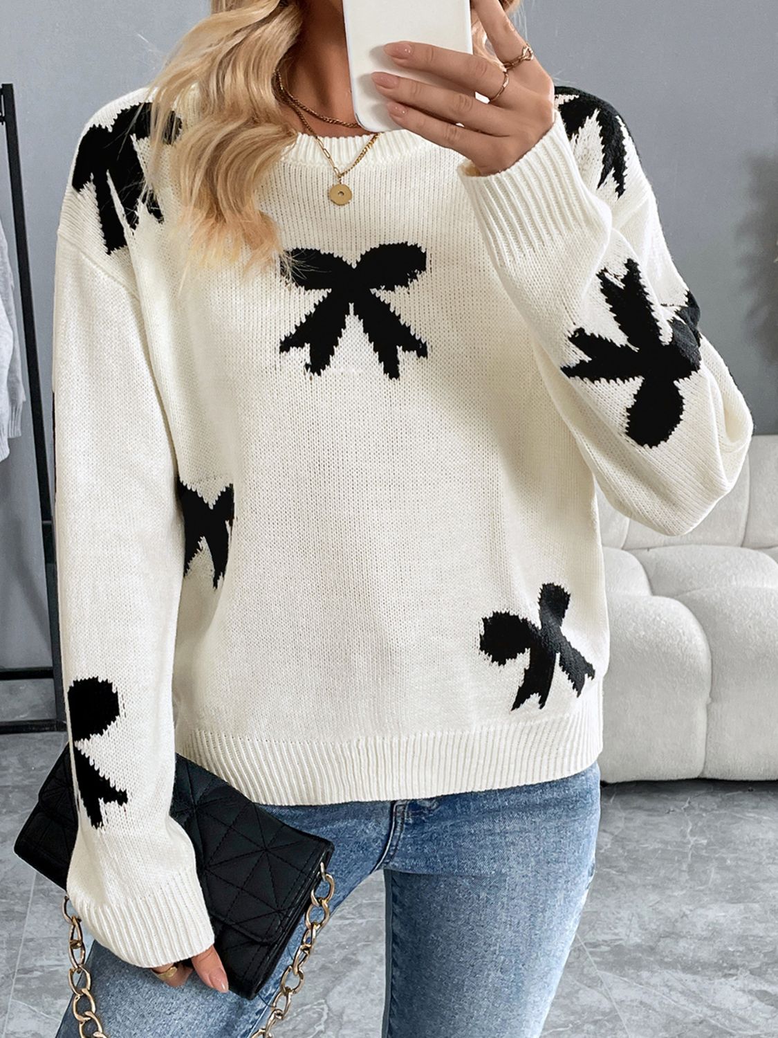 Woman wearing a Perfee Bow Graphic Round Neck Long Sleeve Sweater in white with black bow designs, paired with blue jeans and a black clutch.