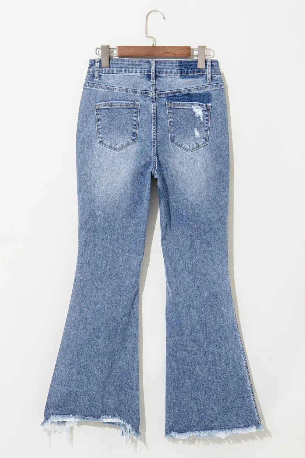 Distressed raw hem bootcut jeans with slightly stretchy material and classic five-pocket design hanging on wooden hanger, featuring back view.