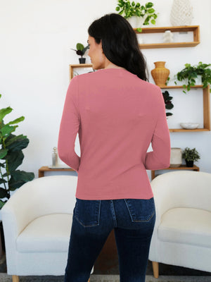 Woman wearing a Dusty Pink ruched mock neck long sleeve t-shirt, standing in a cozy room with plants and white chairs.