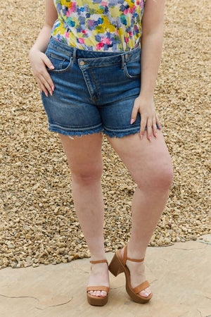 Woman wearing Maya Mid Rise Asymmetrical Shorts by Risen Jeans with colorful top and heeled sandals.