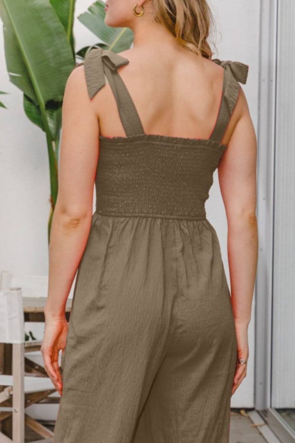 Woman wearing olive green bodice smocked sleeveless jumpsuit with tie straps, back view, standing outdoors.