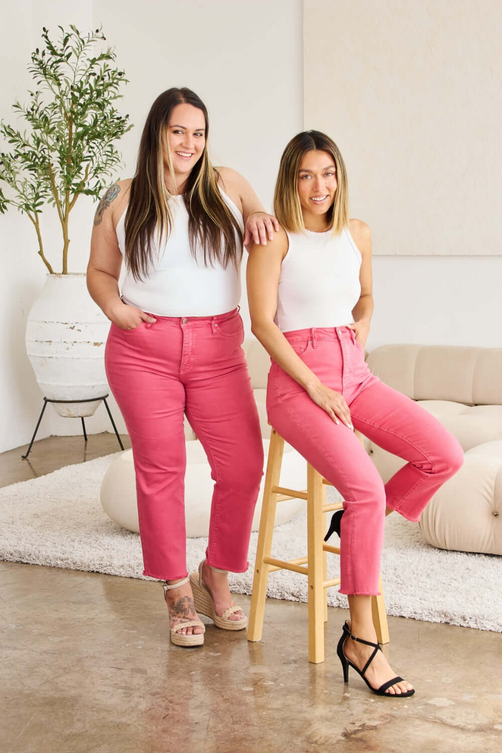 Two women in matching pink RFM Jeans sitting and standing in a stylish room, showing the versatility and comfort of the jeans.