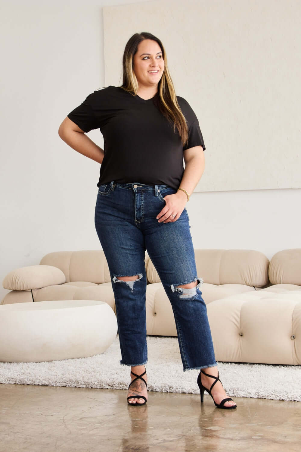 Woman wearing Crop Dylan Full Size Tummy Control Distressed High Waist Raw Hem Jeans by RFM Jeans, standing in a stylish living room.