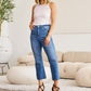 Woman wearing Mini Mia Full Size Tummy Control High Waist Jeans from RFM Jeans, standing in a modern living room.