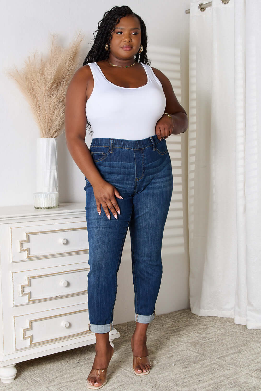 Woman wearing skinny cropped Judy Blue Jeans styled with a white tank top and heels in a modern bedroom.