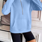 Woman wearing light blue Ivy Lane Half Zip Raglan Sleeve Sweatshirt with front pocket and black shorts, standing by a white wall and decorative items.