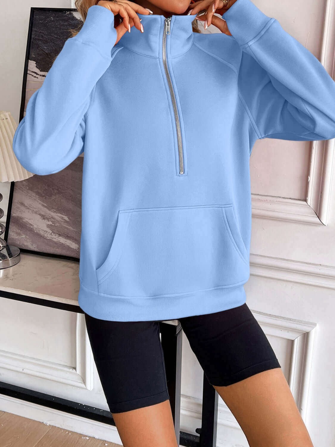 Woman wearing light blue Ivy Lane Half Zip Raglan Sleeve Sweatshirt with front pocket and black shorts, standing by a white wall and decorative items.