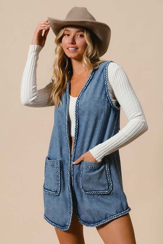 Stylish woman wearing a BiBi braided trim open front denim vest with pockets, paired with a fun hat and long-sleeve top.