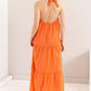 Back view of a woman in an orange halter neck maxi dress with tiered layers, perfect for warm days and tropical getaways.