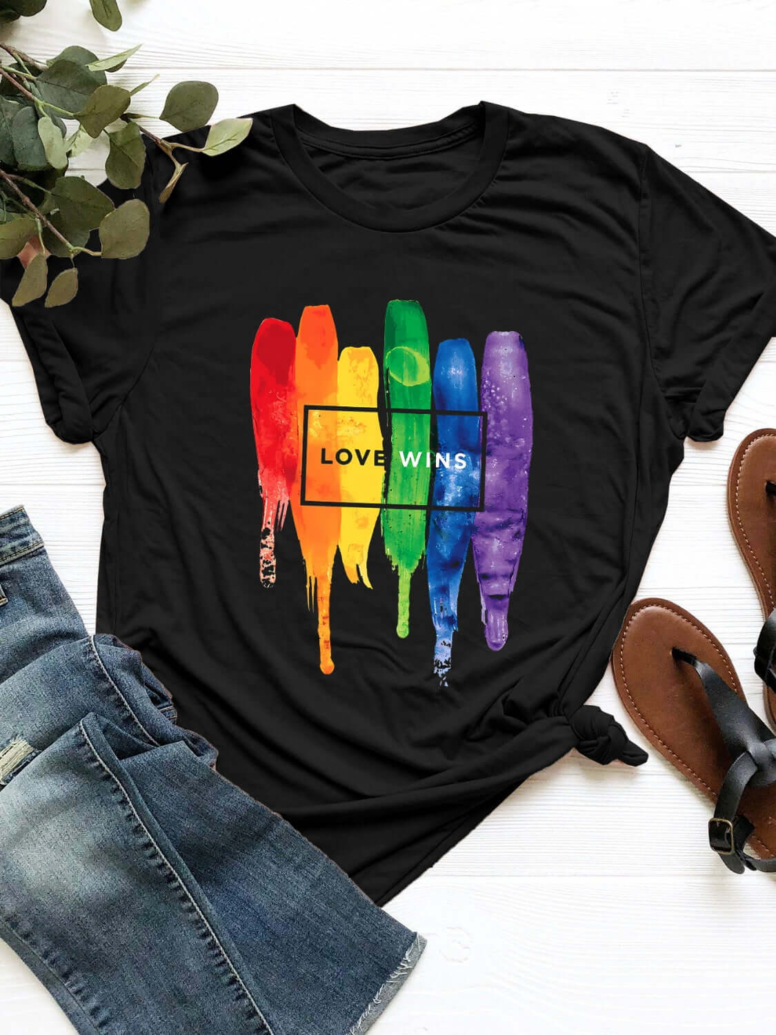 Black graphic round neck short sleeve t-shirt with "Love Wins" rainbow paint stroke design, displayed with jeans and sandals.