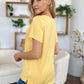 Woman wearing yellow Bamboo V-Neck High-Low T-Shirt, showing back view in a cozy living room setting with green plants and stylish shelves.