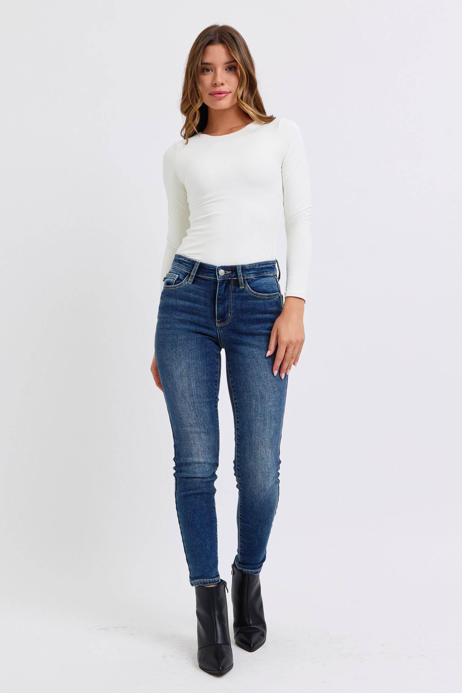 Model wearing Judy Blue mid-rise waist skinny jeans and a white long-sleeve top, showcasing a sleek and stylish look.