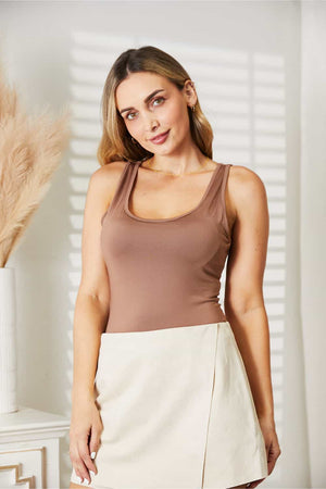 Woman wearing Everyday Full Size Basic Tank Bodysuit in brown, styled with a white skirt, standing in a well-lit room.