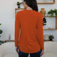 Woman wearing orange FAM-FAM Round Neck Long Sleeve T-Shirt, showcasing back view, in a stylish room with plants and shelves.