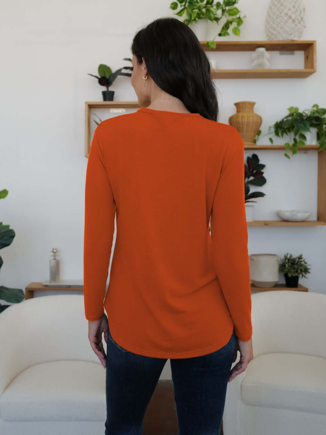 Woman wearing orange FAM-FAM Round Neck Long Sleeve T-Shirt, showcasing back view, in a stylish room with plants and shelves.