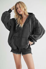 Trendy woman in a black exposed seam zip-up hooded jacket paired with denim shorts, showcasing stylish and casual outerwear.