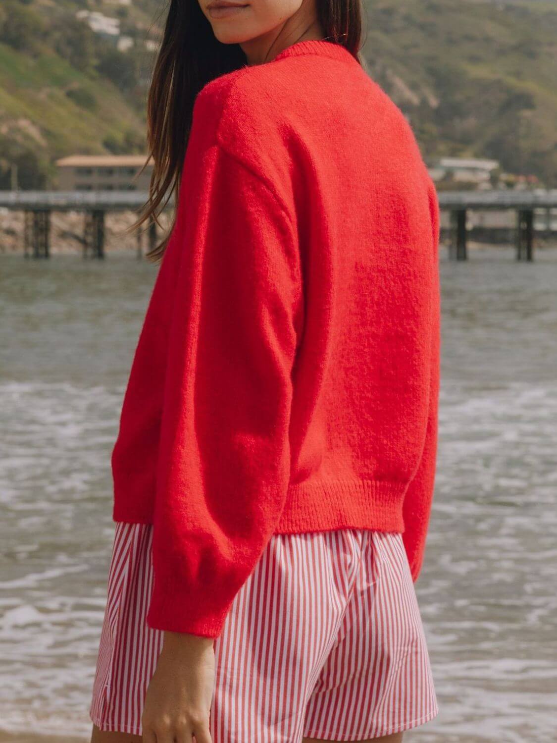 Model wearing a cozy red Bella Road Button Down Cardigan by the beach, paired with striped shorts. Ideal for chilly days!