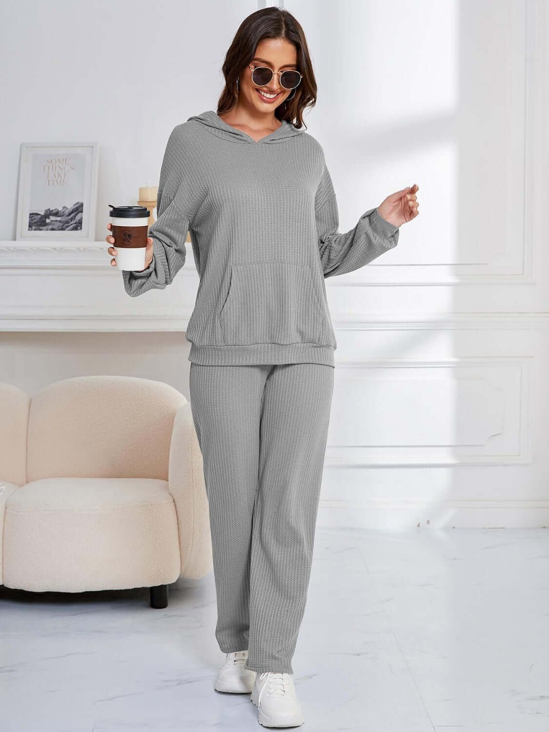 Woman in gray Bella Road hoodie and pants set, holding coffee, in stylish lounge setting.