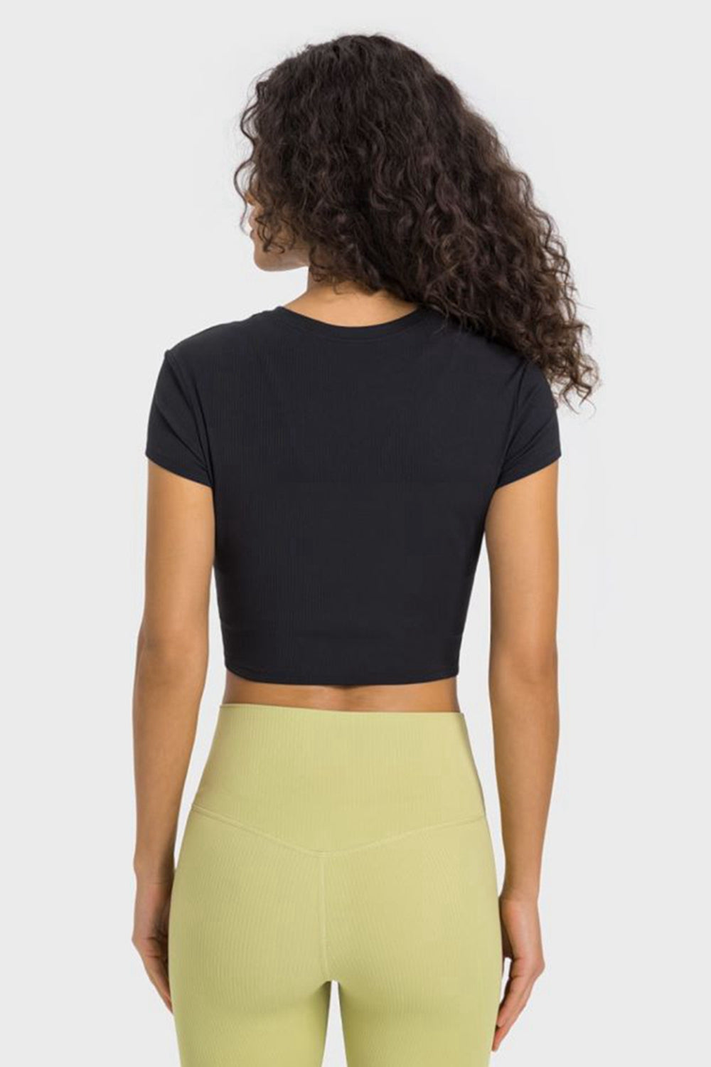 Back view of a woman wearing a black cropped sports t-shirt paired with light green yoga pants. Perfect for workouts!