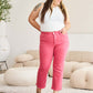 Woman wearing Crop Dylan full size tummy control high waist raw hem jeans in pink, standing in a stylish living room.