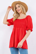 Woman wearing a ruffled short sleeve smocked blouse in red, paired with blue jeans and a wide-brimmed hat.