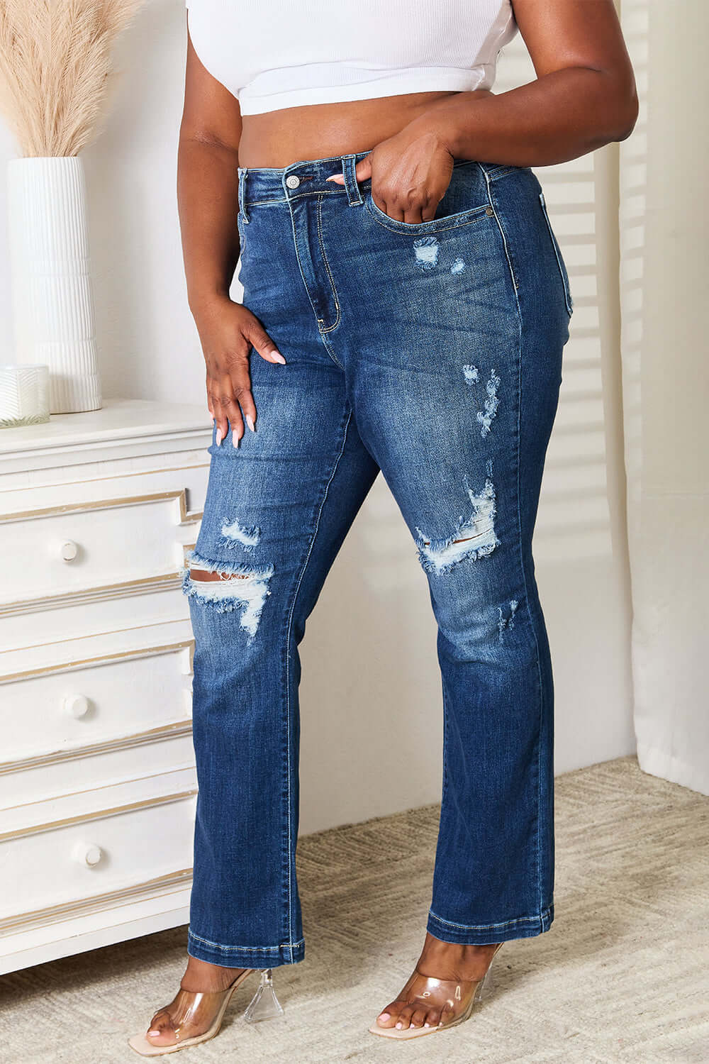 Woman wearing Judy Blue Jeans mid-rise hand sand and destroy bootcut jeans with a distressed look in front of a dresser.