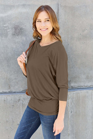 Woman wearing a round neck batwing sleeve top with jeans, standing against a concrete wall.