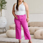 Woman in white tank top and pink high-waisted jeans from RFM Jeans, standing in a stylish living room with a relaxed expression.