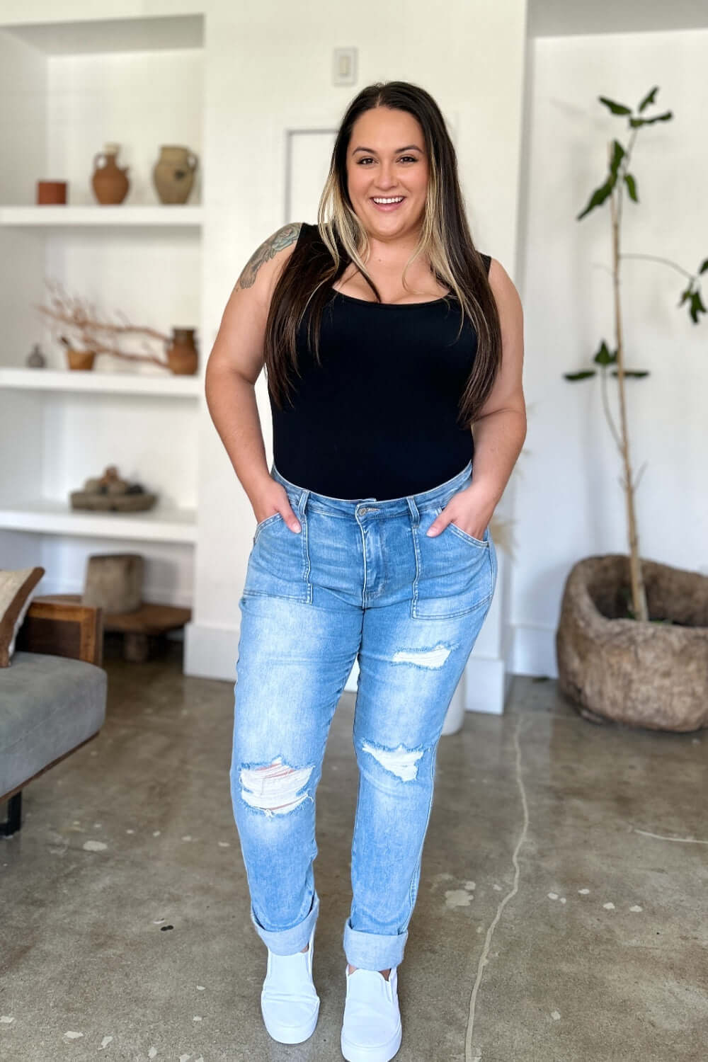 Woman wearing Judy Blue distressed straight jeans with patch pockets, black tank top, standing in minimalist living room.