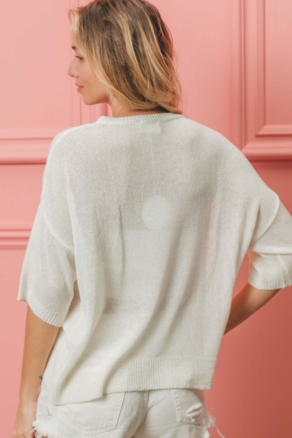 Woman wearing a white knit top with short sleeves and distressed white shorts, standing against a pink paneled wall.