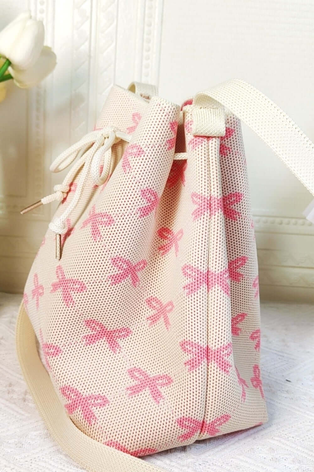 Pink ribbon bowknot pattern crossbody bucket bag with drawstring closure and white strap on a light background.